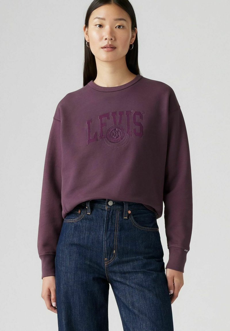 Dunkellila Sweatshirt mit "LEVIS"-Logo in passender Farbe, Rundhalsausschnitt und langen Ärmeln, kombiniert mit hochgeschnittenen dunklen Jeans.
