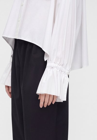 Chemise blanche à manches longues avec poignets froncés et évasés associée à un pantalon noir ample portée par une personne debout devant un fond uni.