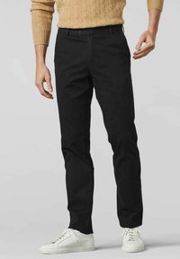 Chino en coton noir avec une coupe slim, présentant une jambe droite, des poches avant et une fermeture à bouton. Porté avec des baskets blanches.