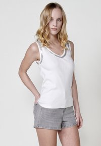 Haut sans manches blanc avec un décolleté en V, présentant des accents décoratifs en argent sur les épaules. Porté avec un short en denim gris. Texture lisse.