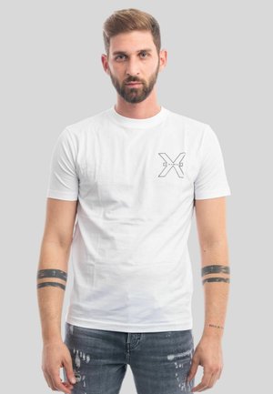 T-shirt bianco in cotone con un design grafico nero di linee intersecting sul lato sinistro del petto. Abbinato a jeans grigi strappati.