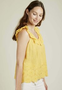 Blouse jaune avec des bretelles à volants, broderies florales et ourlet à volants. Fabriquée en tissu léger, présentant des détails de texture subtils.
