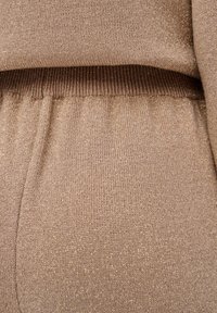 Gebreide trui in beige met subtiele gouden draad, voorzien van een geribde tailleband en een zachte textuur. Close-up gericht op de stofdetails.