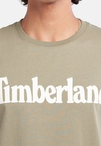 Oliven grøn bomulds t-shirt med rund halsudskæring, der har store hvide "Timberland"-bogstaver på forsiden. Subtil tekstur synlig.