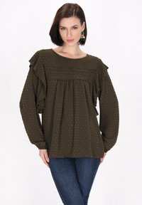 Oliwkowa bluzka z falbaniastymi rękawami, okrągłym dekoltem i teksturowanymi kropkami. Posiada dekoracyjne przeszycia i elastyczne mankiety. Noszona z niebieskimi dżinsami.