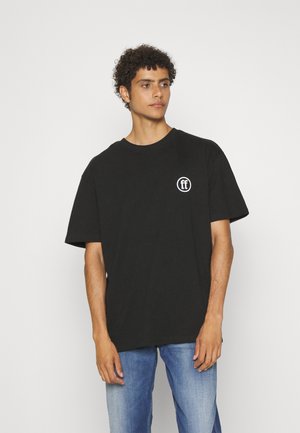 Svart bomull T-skjorte med korte ermer og rund hals, med en hvit logo på venstre side. Brukt sammen med blå denimjeans.