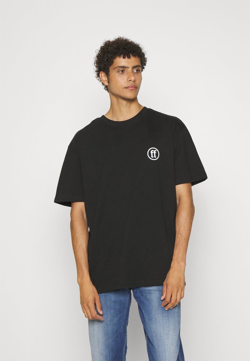 Schwarzes Baumwoll-T-Shirt mit kurzen Ärmeln und rundem Halsausschnitt, mit einem weißen Logo auf der linken Seite. Kombiniert mit blauen Jeans.