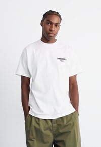 Witte katoenen T-shirt met korte mouwen en een ronde hals, met zwarte tekst op de linkerborst. Gekozen in combinatie met beige, losse broek.