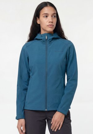 Vrouw met een teal-gekleurde capuchontrui met rits en zwarte broek, staand en licht naar links kijkend tegen een witte achtergrond.