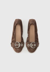 Brune ruskindsflade sko med en rund sølvspænde på remmen, der har et blødt, slip-on design og en glat inderside.