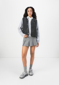 Žena s kudrnatými vlasy, oblečená v šedém kardiganu s copánkovým vzorem, černé fleecové vestě, šedé plisované mini sukni, šedých ponožkách a šedých sportovních botách, stojí.