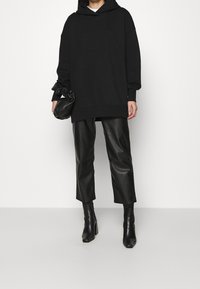 Sudadera negra de gran tamaño con capucha, combinada con pantalones de cuero negros recortados y botas de tacón afilado, sostenida con un pequeño bolso negro de mano.
