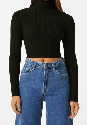 Femme portant un col roulé court noir côtelé à manches longues, associé à un jean bleu en denim taille haute, les mains détendues le long du corps.