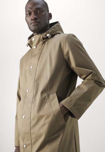 Uomo che indossa un impermeabile color cammello con cappuccio e bottoni a pressione, con una mano in tasca, che guarda leggermente verso il basso su uno sfondo chiaro e uniforme.