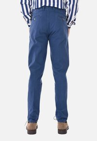 Pantaloni di cotone blu con polsini elastici, caratterizzati da due tasche posteriori con chiusura a bottone e una texture liscia. Abbinati a scarpe marroni.