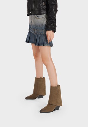 Donna con stivali in camoscio marrone a metà polpaccio, minigonna di jeans plissettata con dettagli di cintura e giacca di pelle nera, in piedi su sfondo semplice.