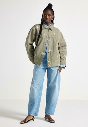 Giacca verde oliva con ricamo floreale, vestibilità oversize, abbinata a jeans a gamba larga azzurri chiaro e sandali neri. Dettagli del tessuto lavorato visibili.
