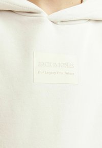 Sudadera gris claro con una tela texturizada, que presenta una etiqueta prominente en beige claro grabada con "JACK & JONES" y texto debajo.
