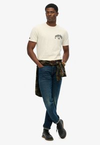 Beige short-sleeve t-shirt with black graphic print, paired with blue jeans. A plaid shirt tied around the waist and black boots complete the look.