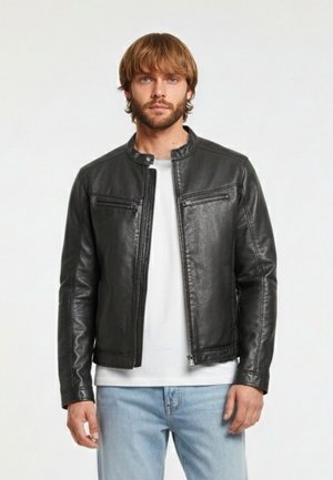 Mann mit hellbraunen Haaren und Bart, trägt eine schwarze Lederjacke über einem weißen Hemd und hellblauen Jeans, steht vor einfachem Hintergrund.