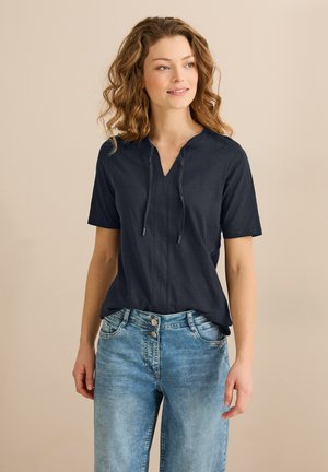 Top de manga corta azul marino con escote en V, que presenta un ligero fruncido en el cuello y un corte relajado. Combinado con jeans de mezclilla azul claro.