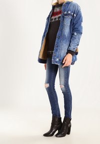 Chaqueta de denim, azul con detalles desgastados, superpuesta a un suéter negro con patrones en rojo y gris. Jeans ajustados con rasgaduras en las rodillas y botas negras hasta el tobillo.
