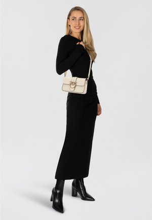 Mala branca estruturada com detalhes pretos e ferragens douradas, usada em cruzado. Modelo com vestido preto justo de manga comprida e botas de salto alto.