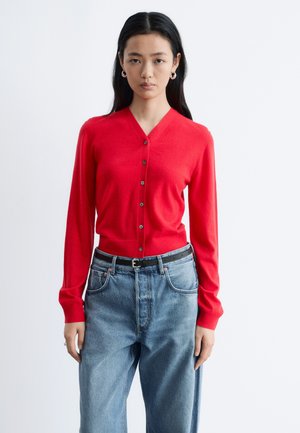 Jonge vrouw die een rood vest met knopen draagt en een blauwe spijkerbroek met een zwarte riem, staand tegen een effen witte achtergrond.