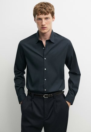 Ung mand iført en tætsiddende sort skjorte med knapper og sorte bukser med et læderbælte, stående mod en ensartet lys baggrund.