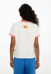 Osoba s dlouhými kudrnatými vlasy má na sobě bílé tričko s růžovým lemem a malou barevnou grafikou na horní části zad, spárované s modrými kalhotami.