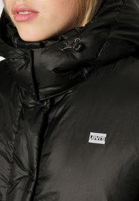 Doudoune noire avec un col haut, arborant un patch logo Levi's visible et des fermetures à boutons. Le tissu présente une finition brillante et texturée.