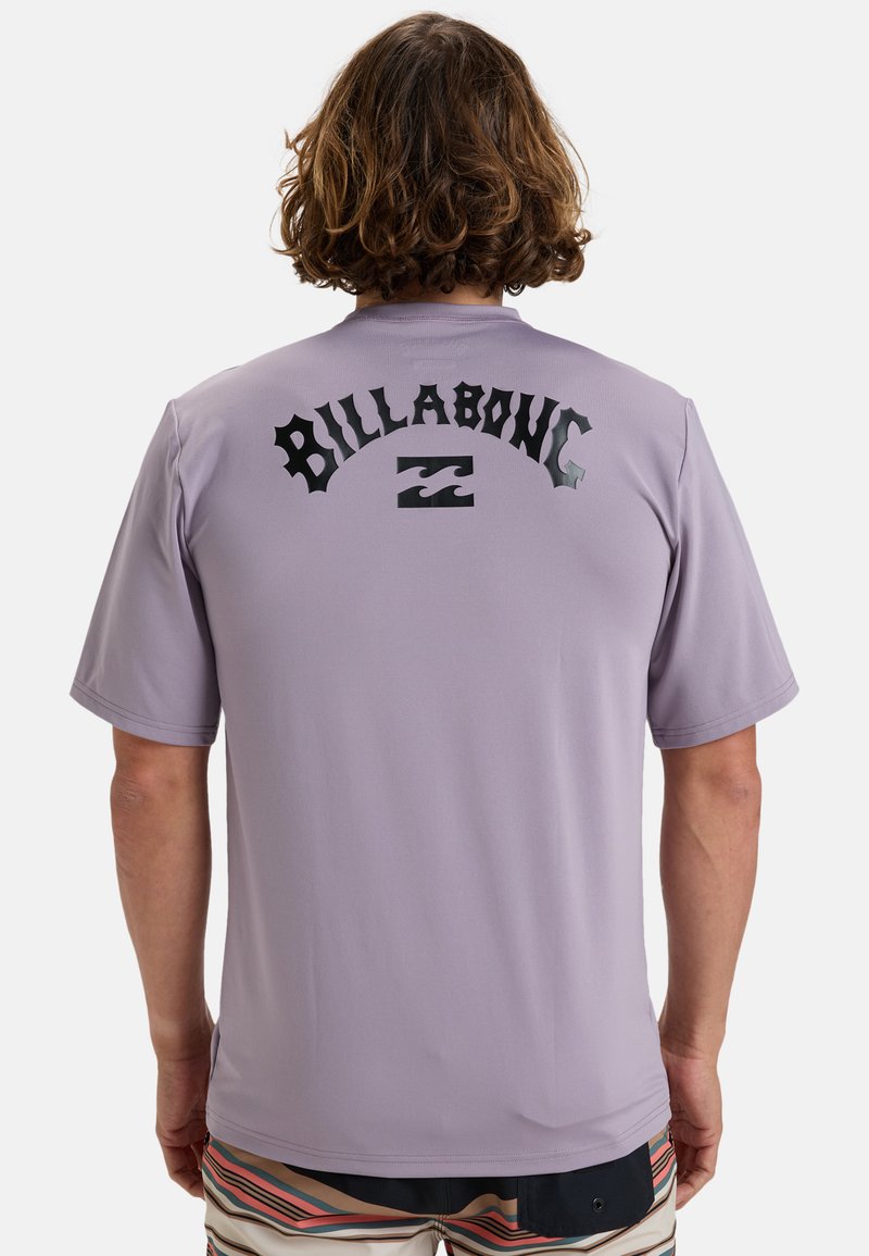 Camisa de manga corta morada con un gráfico negro de "Billabong" en una fuente en negrita en la parte posterior, fabricada con un material suave y ligero.