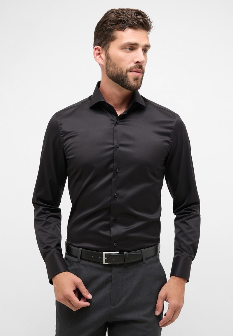 Camicia nera a maniche lunghe realizzata in tessuto liscio, con colletto, chiusura con bottoni e taglio aderente. Abbinata a pantaloni grigi e cintura.
