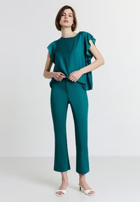 Donna che indossa un top con maniche arricciate in teal e pantaloni cropped abbinati, con sandali aperti bianchi con tacco, in piedi di fronte a uno sfondo neutro.