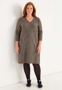 Klänning med V-ringning och lång ärm i ett texturerat, dämpat brunt tyg; kombinerad med svarta strumpbyxor och sneakers för en avslappnad stil.
