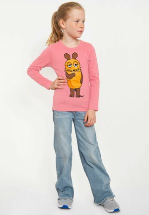 Junges Mädchen mit blondem Pferdeschwanz, trägt ein rosa Langarmshirt mit einem Cartoon-Maulwurf, blaue Jeans und graue Turnschuhe, steht mit der Hand in der Hüfte.