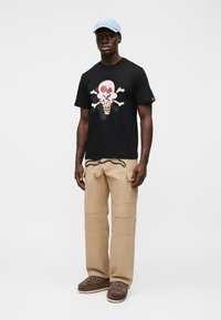 Μαύρο T-shirt με πολύχρωμο σχέδιο παγωτού και κόκκαλα. Μπεζ παντελόνι cargo με τσέπες, καφέ παντόφλες με ανθισμένα σχέδια. Ανοιχτό μπλε καπέλο.