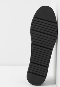 Semelle de chaussure noire avec des rainures horizontales antidérapantes et le numéro de taille 37 centré, positionnée debout sur un fond blanc.