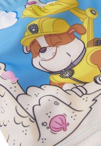 Costume da bagno colorati con un cane dei cartoni animati che indossa un casco giallo, sorridente su uno sfondo blu con sabbia e conchiglie.