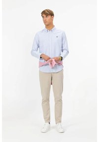 Giovane uomo che indossa una camicia a righe azzurre chiusa con bottoni e pantaloni beige, che si lega un maglione rosa intorno alla vita, in piedi su sfondo bianco.