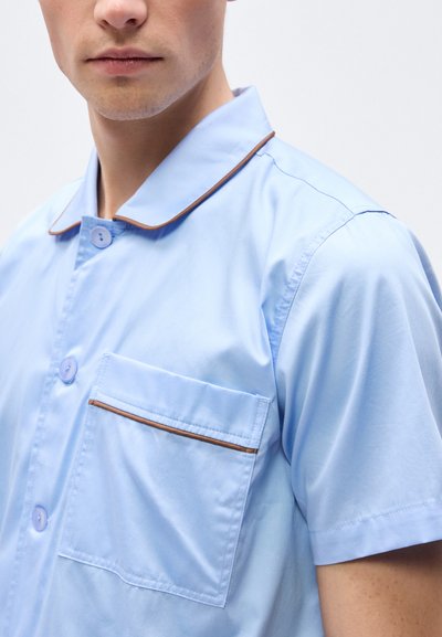 Άνδρας που φοράει γαλάζιο κοντομάνικο πουκάμισο με καφέ περιθώριο στο γιακά και την τσέπη, κουμπωμένο, με φόντο απλό και φωτεινό.