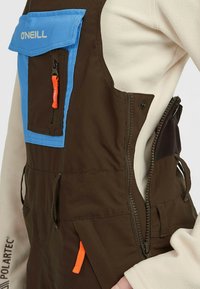 Bruine overall met blauwe accenten, voorzien van een grote blauwe borstzak en een oranje ritssluiting. Gemaakt van duurzame stof, met een taps toelopende taille.