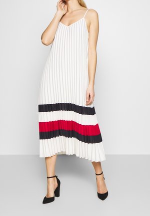 Femme portant une robe midi plissée blanche sans manches avec des rayures verticales et des blocs de couleur noirs, blancs et rouges horizontaux près de l'ourlet.