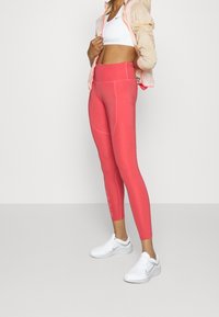 Leggings roses à taille haute avec une texture lisse et des détails de couture, associés à un soutien-gorge de sport blanc et une légère veste beige.