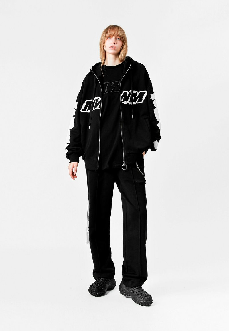 Sudadera negra con cremallera y logo blanco en las mangas, combinada con una camiseta negra y pantalones negros holgados. Unos zapatos negros texturizados completan el conjunto.
