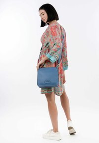 Borsa a tracolla in pelle blu con finitura testurizzata, forma rettangolare e tracolla regolabile, indossata insieme a un vestito a fantasia e sneakers bianche.