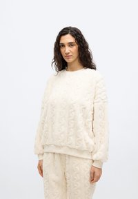 Γυναίκα με καστανά σγουρά μαλλιά, που φοράει ένα κρεμ υφασμάτινο φλις φούτερ και ταιριαστό παντελόνι, στέκεται μπροστά σε ένα απλό φωτεινό φόντο.