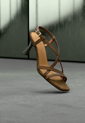 Καφέ δερμάτινο πέδιλο με λουράκια και τακούνι, με λεία υφή, σχέδιο ανοιχτών δαχτύλων, αγκράφα στον αστράγαλο και κομψό μαύρο τακούνι σε μια μινιμάλ φόντο.