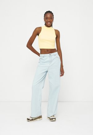 Jeune femme souriante, debout avec une main sur la hanche, portant un haut court sans manches jaune, un jean large bleu clair et des baskets.