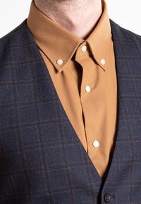 Marineblauer Karo-Weste mit einem braunen Hemd mit Kragen darunter. Das Hemd hat weiße Knöpfe und eine glatte Textur. Die Weste hat ein weiches Finish.